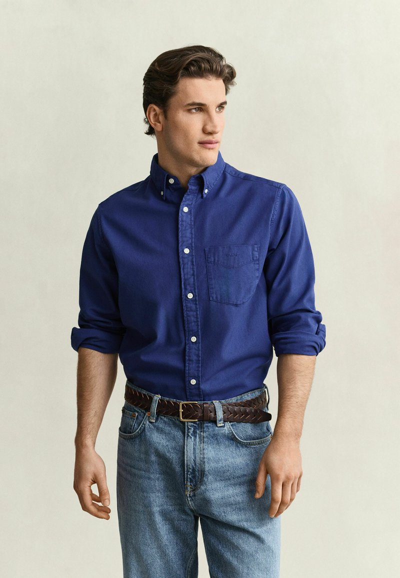 Junger Mann mit blauem Hemd mit Knöpfen und hochgekrempelten Ärmeln, hellblauen Jeans und braunem geflochtenem Gürtel, der nach rechts schaut.