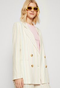 Dubbelbijde blazer in lichte pastel gestreepte stof met gouden knopen, gecombineerd met een roze top en gele zonnebril. Zachte textuur.