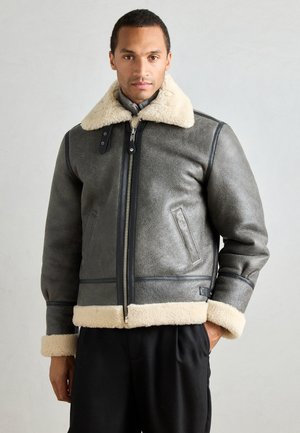 Grå läderjacka med shearlingfoder, mjuk krämfärgad krage och manschetter, med en framdragkedja och sidofickor. Ren, strukturerad design.