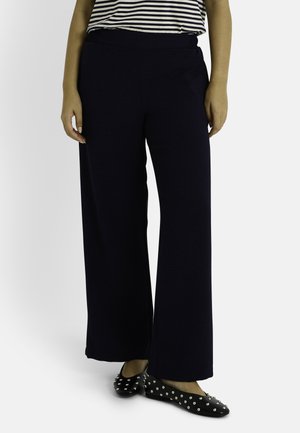 Pantalons larges, bleu marine foncé avec une taille élastique, fabriqués en tissu texturé. Associés à des ballerines noires avec des clous argentés.
