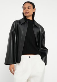 Sort læderjakke med krave, der har knaplukning og vide ærmer, båret over en sort ribstrikket sweater og hvide bukser.