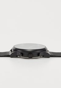 Montre chronographe noire avec lunette lisse, boîtier rond, bracelet texturé et trois boutons latéraux ; présente un cadran minimaliste avec des accents verts.