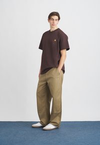 Dark brown cotton t-shirt with short sleeves and a small logo, paired with loose, khaki-colored pants and white athletic shoes.
