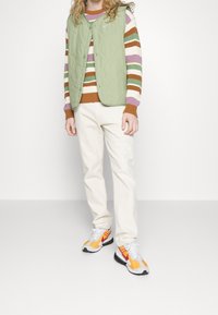 Person i grön vadderad väst, randig flerfärgad tröja, off-white byxor och orange-vita sneakers står på en enkel bakgrund.