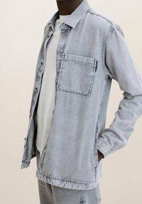 Camicia in denim grigio chiaro con design a bottoni, taschino sul petto, maniche lunghe e una leggera trama. Indossata sopra una maglietta bianca.