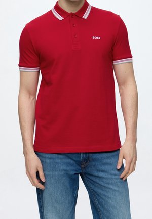 Mand iført en rød polo med hvid kant på krave og ærmer, broderet BOSS-logo på brystet og blå jeans.