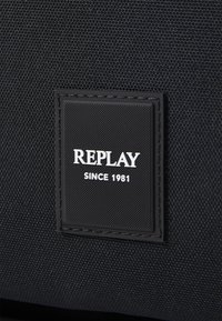 Bolsa de tela negra con una superficie texturizada y una etiqueta rectangular con el texto "REPLAY SINCE 1981" en blanco.