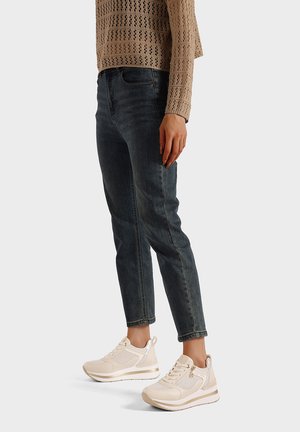 Persona con jeans blu scuro, maglia beige a maniche lunghe traforata e sneaker bianche con plateau, in piedi contro uno sfondo semplice.