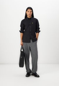 Camicia nera con bottone, logo giallo, pantaloni a scacchi grigi, scarpe nere e borsa tote nera. Il modello è in piedi davanti a uno sfondo bianco.