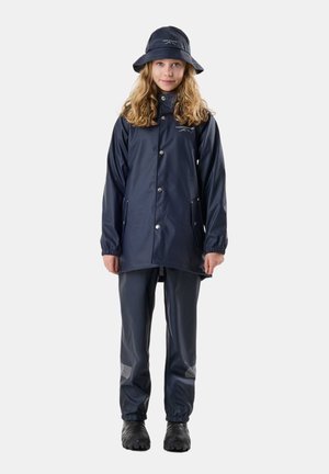 Kind met krullend haar, gekleed in een marineblauwe regenjas, bijpassende broek met reflecterende stukken, regenlaarzen en een vissershoed, staand tegen een witte achtergrond.