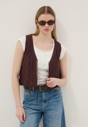 Junge Frau trägt Schildpatt-Sonnenbrille, weißes Kurzarm-Top mit gewellter Spitze, braune Strickweste, blaue Jeans und Ledergürtel.