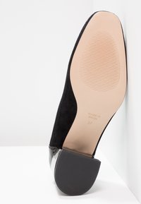 Chaussure femme en daim noir inclinée montrant une semelle beige avec grip rainuré, talon bloc, taille 37, fabriquée en Espagne, texte visible.