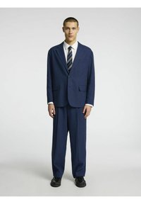 Costume bleu marine avec une coupe décontractée, comprenant un blazer à un bouton et un pantalon évasé. Associé à une chemise blanche et une cravate rayée.
