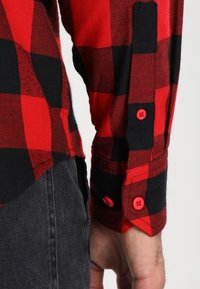 Mouw van een rood en zwart geruite flanellen shirt met rode knopen, die rust naast een hand boven donkergrijze broek.