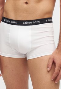 Homme portant un boxer blanc avec une ceinture noire de marque Björn Borg, montrant le torse et le haut des cuisses sur fond blanc.