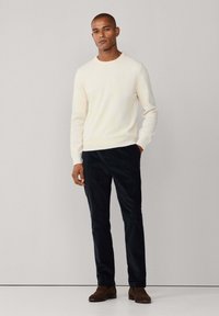 Cremfarbener Strickpullover mit Rundhalsausschnitt, kombiniert mit dunkelblauen Cordhosen und braunen Wildlederschuhen, vor einem neutralen Hintergrund stehend.