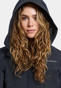 Young woman with long curly blonde hair wearing a black hooded jacket with the hood up, looking directly ahead.