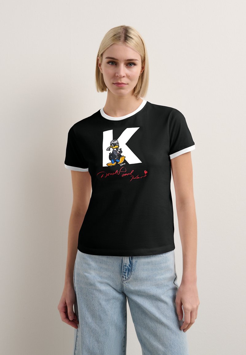 Schwarzes T-Shirt mit kurzen Ärmeln und weißen Abschlüssen, das ein großes weißes 'K' und eine Grafik eines Cartoon-Charakters auf einem Skateboard zeigt.