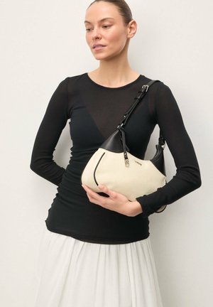 Frau in schwarzem Langarmoberteil und weißem Rock hält eine beige-schwarze Handtasche mit Schulterriemen vor einem einfarbigen Hintergrund.