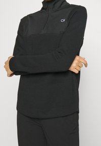 Pull en polaire noir zippé avec une partie supérieure texturée et une partie inférieure lisse. Présente le logo CK sur la poitrine gauche et des poignets ajustés.