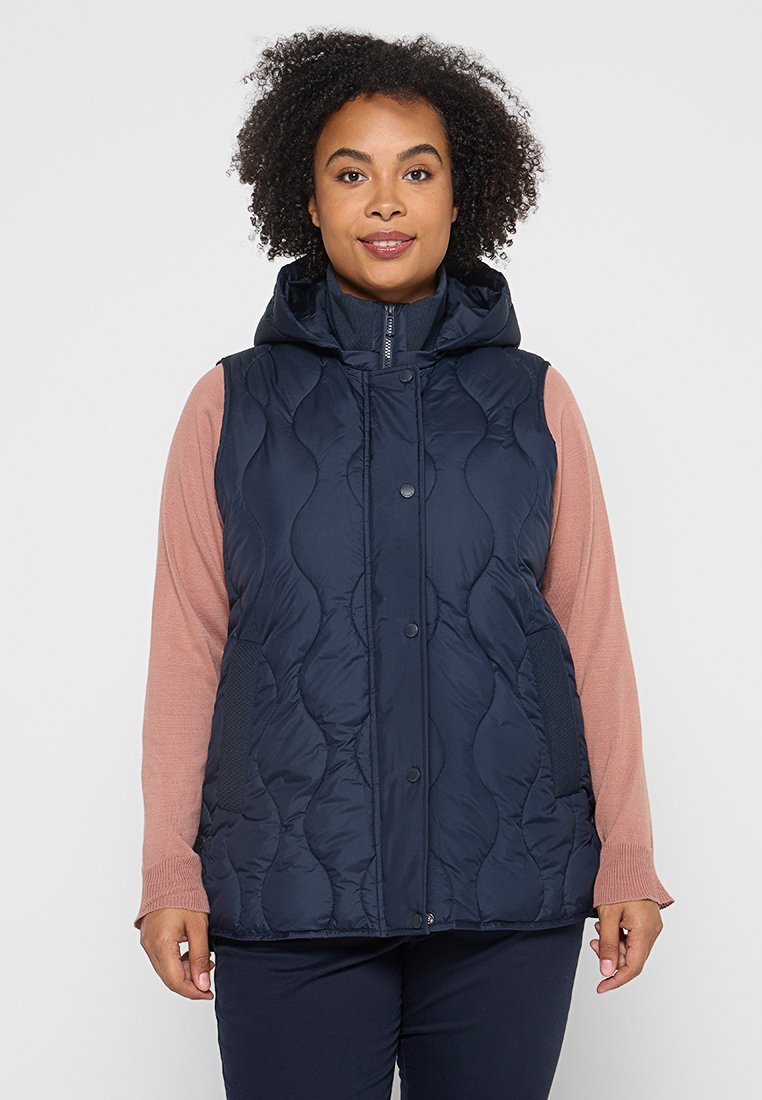 Gilet matelassé bleu marine avec une capuche, présentant un motif matelassé et une fermeture à boutons-pression. Porté par-dessus un haut à manches longues rose clair.