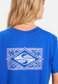 Blaues Baumwoll-T-Shirt mit Rundhalsausschnitt, mit einem diamantförmigen Grafikdesign in Weiß und Pink auf der Rückseite, das ein strukturiertes Design zeigt.