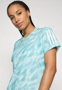 Giovane donna con capelli intrecciati che indossa una maglietta sportiva Adidas a righe blu chiaro con motivi e strisce bianche sulle maniche.