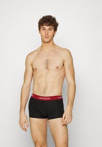 Fekete pamut boxer alsónadrág piros derékrésszel, amelyen a "Lacoste" logó található. A dizájn testhez simuló és sima, minimális varrásokkal.