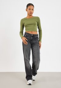 Zöld bordázott, hosszú ujjú crop top fekete egyenes szárú farmernadrággal párosítva. A modellen világosszürke sportcipők vannak. Minimalista dizájn, semleges háttér.