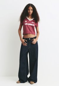 Ung kvinde med krøllet hår iført en maroon cropped "Los Angeles" trøje og brede mørkeblå denim jeans står foran en hvid baggrund.