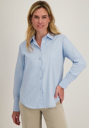 Vrouw met blond haar die een lichtblauw overhemd met knopen en beige broek draagt, staand met één hand op haar heup tegen een neutrale achtergrond.