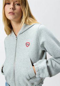 Felpa con cappuccio grigia chiara con zip e toppa a forma di cuore. Presenta una zip in metallo, tasche laterali e polsini a coste. Tessuto morbido con una texture liscia.