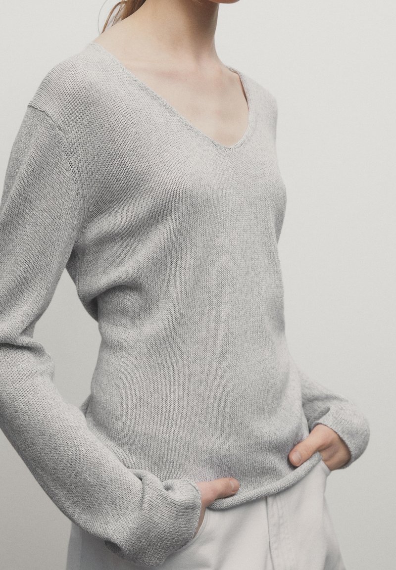 Femme portant un pull maille gris clair à col en V avec manches longues, mains dans les poches d'un pantalon blanc, sur un fond uni.