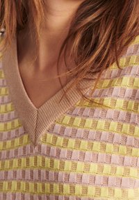 Nærbillede af en person iført en gul og lys lilla ternet striktrøje med en beige V-hals krave og brunt hår.