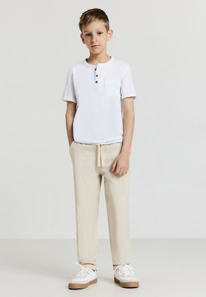 Weißes kurzärmeliges Henley-Shirt mit einer Tasche und Knöpfen, kombiniert mit hellbeigen Jogginghosen mit Kordelzug und weißen Sneakers mit brauner Sohle.
