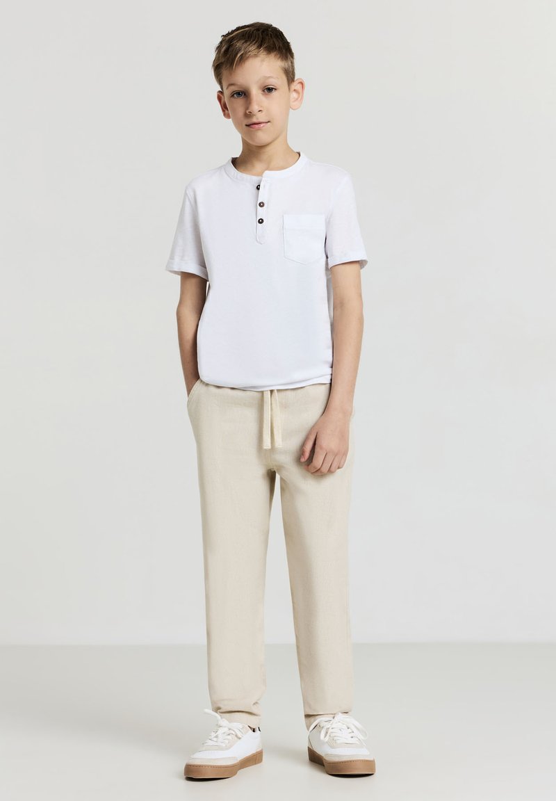 Weißes kurzärmeliges Henley-Shirt mit einer Tasche und Knöpfen, kombiniert mit hellbeigen Jogginghosen mit Kordelzug und weißen Sneakers mit brauner Sohle.
