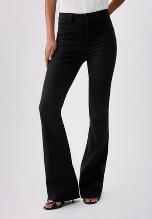 Kobieta ubrana w czarne spodnie typu flare z wysokim stanem i naszywanymi kieszeniami, zestawione z czarnymi sandałami na obcasie z odkrytymi palcami i białą bluzką, stojąca na jednolitym tle.