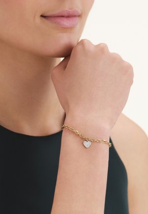 Woman wearing a gold chain bracelet with a small heart-shaped charm covered in diamonds on her left wrist, hand near face.