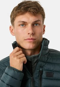 Giacca puffer verde scuro con colletto alto, chiusura con zip e texture trapuntata. Presenta una toppa con logo nera sul petto.