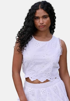 Žena s dlhými kučeravými čiernymi vlasmi, ktorá nosí biely vyšívaný crop top bez rukávov a zodpovedajúcu sukňu, s neutrálne výrazom a svetlým pozadím.