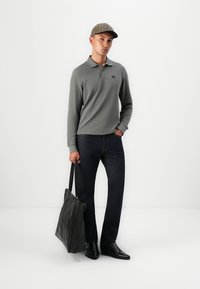 Grau langärmliges Polo-Shirt mit Logo, dunkle Jeans, schwarze Leder-Tasche und eine Kappe mit Karomuster. Mann steht vor einem schlichten Hintergrund.