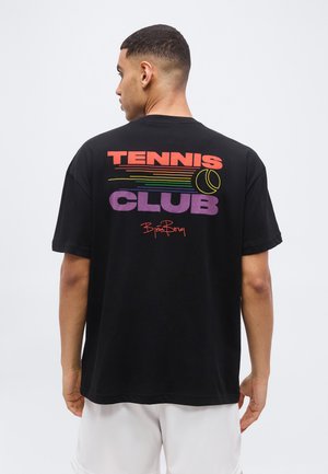Άνδρας με μαύρο T-shirt που γράφει "TENNIS CLUB", πολύχρωμες γραμμές και γραφικό με μπάλα του τένις στην πλάτη, κοιτάζοντας προς τα πίσω.