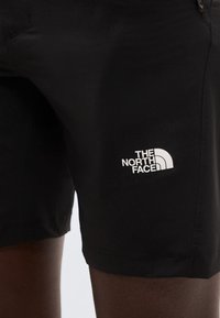 Lähikuva mustista shortseista, joissa on valkoinen The North Face -logo etureidessä, henkilö jolla on tumma iho ja jotka ovat päällä.