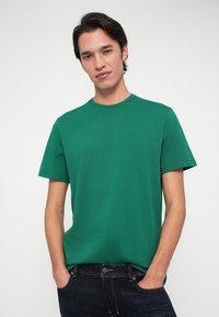 Jaunas vyras su tamsiais plaukais, vilkintis žalią trumpomis rankovėmis marškinėlį ir tamsias džinsus, su rankomis kišenėse, stovi prieš paprastą šviesią fono.