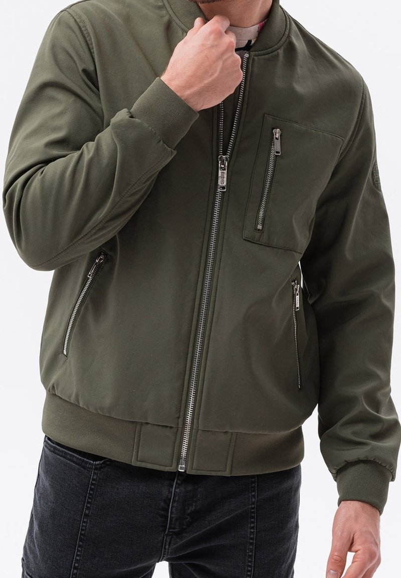 Homme portant une veste bomber vert olive avec plusieurs poches zippées et un jean foncé, ajustant le col d'une main.