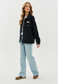 Czarna kurtka dżinsowa z dużymi kieszeniami z przodu, jasnoniebieskie rozkloszowane dżinsy i brązowe buty wsuwane w panterkę. Modelka stoi z profilu.