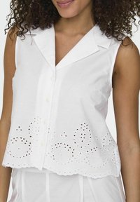 Blusa bianca senza maniche con colletto, chiusura frontale a bottoni e ricamo decorativo a occhio lungo l'orlo.