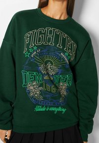Grön sweatshirt med framsidan grafisk design med "FIGHTED," berg, blommor, och "DENVER," accentuerad med guld- och blå sömnad.