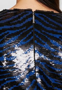 Gros plan sur un tissu à sequins bleu, noir et argenté avec une fermeture éclair visible au centre du dos d'un vêtement porté par une personne.