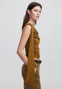 Top mostaza sin mangas con diseño drapeado y una larga banda de hombro fluida. Combinado con pantalones marrones texturizados y un cinturón trenzado.